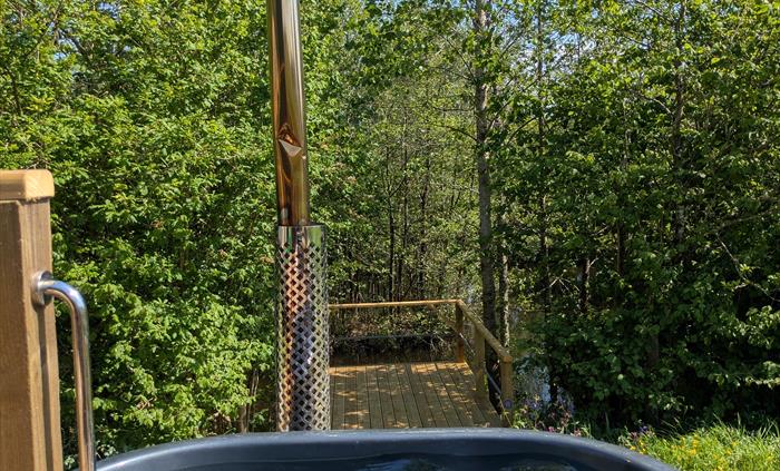Grey fibreglass hot tub in wood casing next to the wood deck, with a metal chimney behind, overlooking the lake and trees in the background. There are